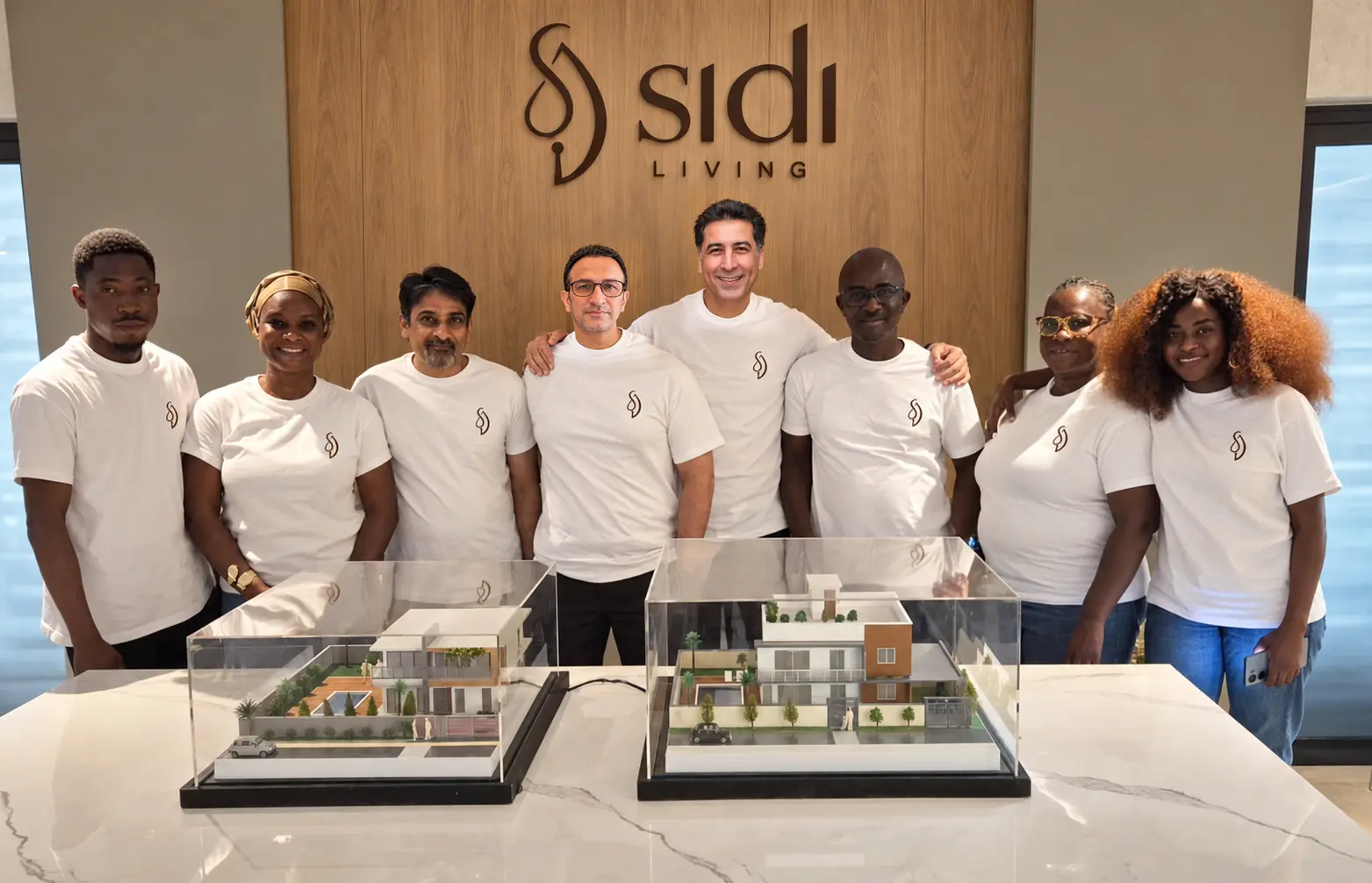 The SIDI Living team standing beneath the SIDI Living wall logo with architectural models in front
